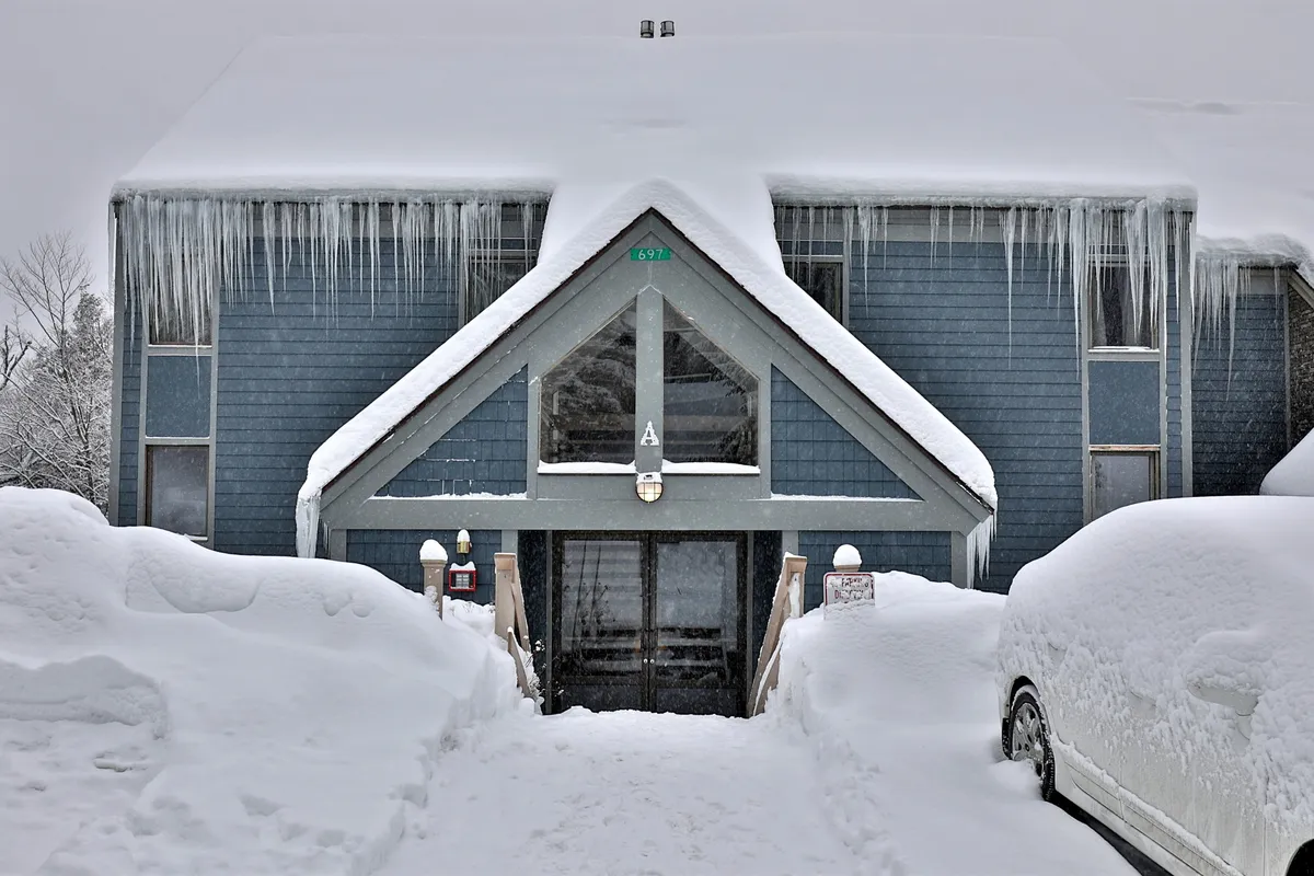 A close-up of the snow-covered entrance to the condo, with icicles dangling from the roof and wooden steps leading to the front door. The setting feels serene and quietly inviting, suggesting a welcoming refuge after outdoor activity.