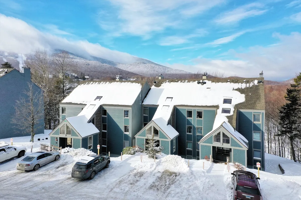 A snow-covered condo complex nestled against a stunning mountain backdrop. The roofs and parking area are blanketed in fresh white snow, evoking the heart of ski season. The scene captures the proximity to outdoor adventure and the inviting, cozy refuge awaiting after a day on the slopes.