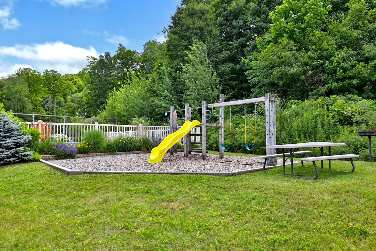 A charming outdoor shared space featuring playground equipment, a vibrant yellow slide, a swing set, and a picnic table nestled amidst lush greenery. Families and friends gather for picnics and playful afternoons under the sun.