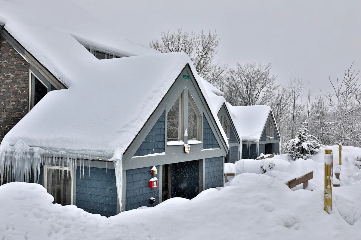 A charming winter exterior of the property, featuring snow-covered roofs, icicles hanging from the eaves, wooden railings, and soft snow drifts all around. The scene invokes the feeling of returning to a cozy mountain base after outdoor adventures.