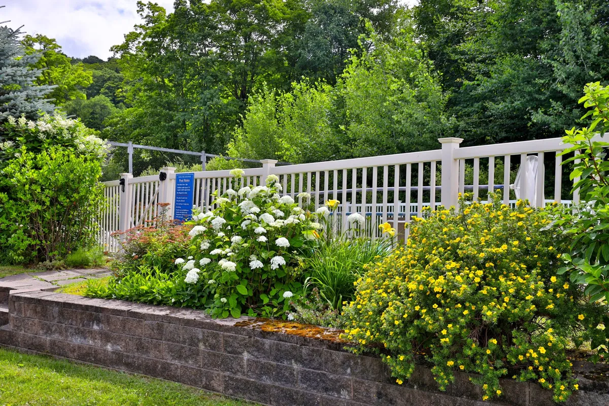 A vibrant exterior scene filled with blooming flower beds, a white picket fence, and abundant green foliage. The space is cheerful and inviting, ideal for family barbecues, picnics, or enjoying a morning coffee outdoors.