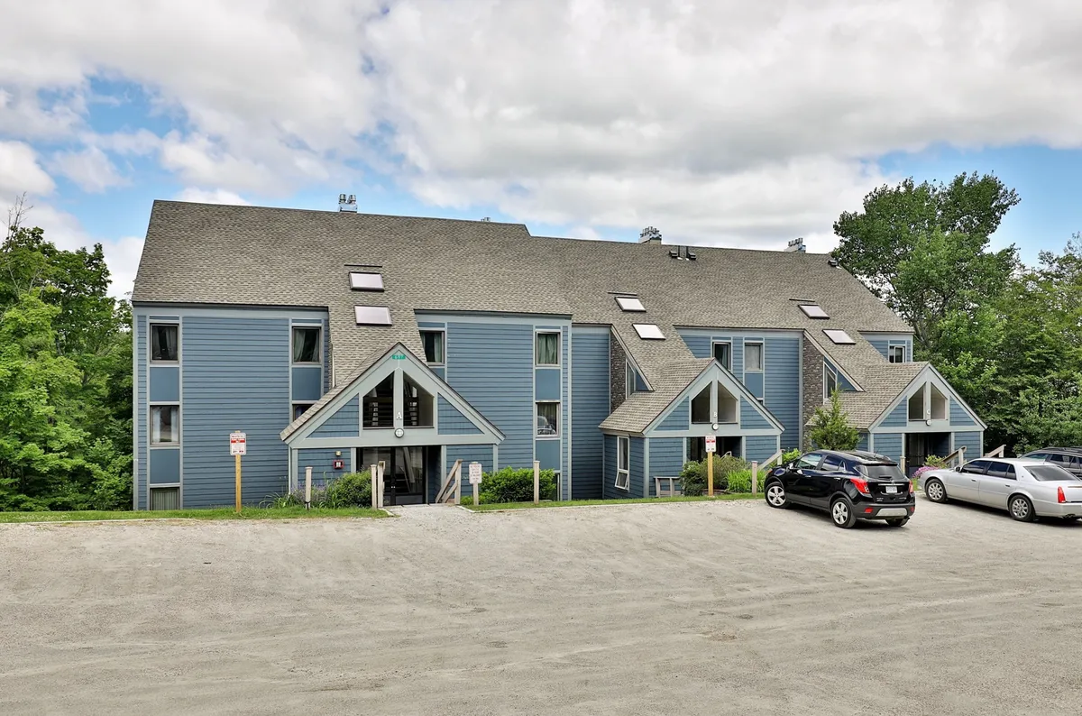 The exterior of the condo complex, displaying blue wooden siding, unique triangular rooflines, windows with skylights, and a tranquil setting framed by greenery. The parking area is visible, suggesting easy accessibility for families.