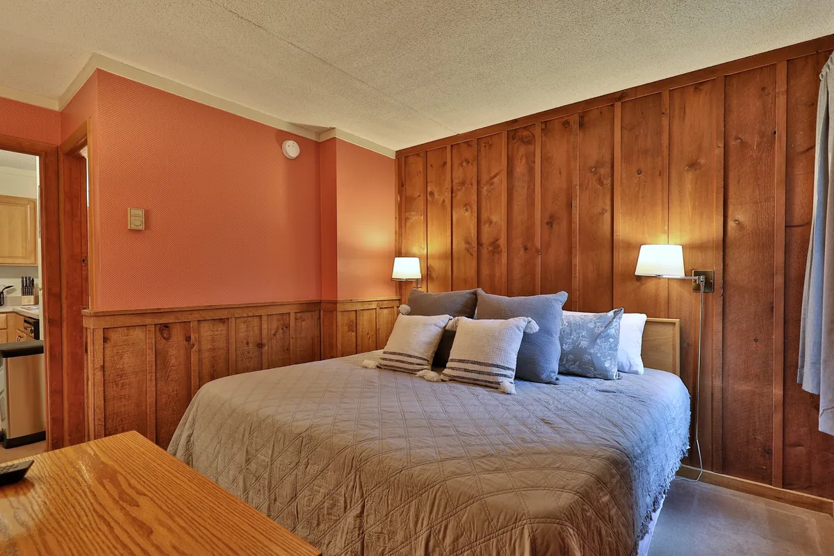 A warm, wood-paneled bedroom featuring a plush bed adorned with decorative pillows and flanked by wall lamps. The atmosphere is cozy, emphasizing relaxation and comfort, ideal for restful nights or quiet mornings.