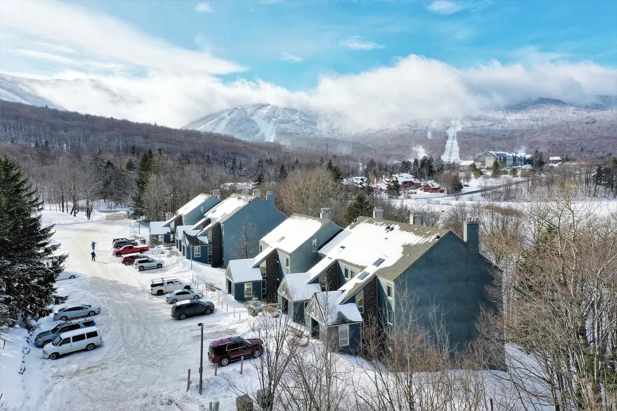 The image showcases the Whiffletree condo complex blanketed in fresh snow, framed by tall trees and set against a breathtaking mountain backdrop. The early morning light catches on the snow-covered rooftops, while a few cars are parked along a plowed path. The overall atmosphere is peaceful and inviting, evoking the promise of winter adventures and cozy indoor evenings.
